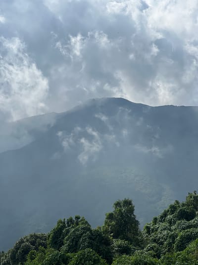 The image depicts a mountainous landscape under a cloudy sky in El Congo, El Salvador. The upper portion of the frame features a densely clouded sky, with patches of bright white cumulus clouds interspersed with darker grey overcast areas, indicating diffused lighting. Below the sky, multiple layers of mountains recede into the distance. The closer mountain ranges are partially obscured by thick mist or low-hanging clouds, creating a hazy, ethereal effect and softening their contours. Distant peaks appear as darker silhouettes against the brighter sky, demonstrating atmospheric perspective. The slopes of these mountains are covered in dense, dark green foliage, visible as textured patterns through the mist. The foreground is dominated by a lush, vibrant canopy of tropical or subtropical trees and thick vegetation. The leaves are a rich green, appearing healthy and forming an intricate texture of overlapping foliage. No human figures, animals, or man-made structures are discernible within the scene. The overall impression is one of a serene and misty natural environment.