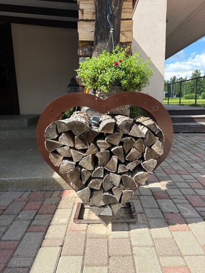This image depicts an outdoor scene, likely in Mežpļavas, Latvia, featuring a heart-shaped metal rack filled with neatly stacked firewood. The firewood is prominently displayed in the foreground, occupying the central and lower portions of the frame. Behind the firewood rack, a portion of a building is visible, characterized by a light-colored, textured wall and a dark-colored roof overhang. To the left of the building, there are concrete steps leading up to an entrance. A hanging lantern and a lush green plant with small red flowers are placed near the building, adding decorative elements. The ground is paved with interlocking bricks in shades of grey, red, and beige. In the background, a wooden deck, a body of water, and trees are visible under a bright blue sky with scattered clouds, suggesting a natural or rural setting adjacent to the urban area. There are no people visible in the image.