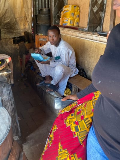 An indoor or semi-enclosed commercial or residential space in Sabon Gari South, Nigeria, is depicted. In the foreground, partially visible on the right, is a person wearing a black long-sleeved top, blue jeans, and a red and yellow patterned fabric wrapper. This individual holds a silver-colored smartphone, directed downwards.
Centered in the midground, a young male, appearing to be in his late teens or early twenties, sits on a wooden bench. He is dressed in a light-grey, long-sleeved tunic (agbada) and black slide sandals. He holds a light blue plastic container with food and a spoon, actively engaged in eating. A green-cased mobile phone is visible within his tunic's left pocket. His gaze is directed upwards and to the right of the frame.
The background features rough, possibly corrugated metal or concrete walls. A wooden shelf on the upper wall displays multiple stacked yellow packages labeled "Meander." Below the shelf, stacked dark metal containers and an orange plastic crate are visible. The floor is composed of dark, rectangular tiles or paving stones. On the far left, a large, dark, possibly wooden structure with a circular top is partially visible. Natural light illuminates the scene, casting shadows.