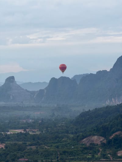An aerial view captures a serene landscape in Ban Phon Sai, Laos, dominated by lush green foliage and towering limestone karsts. The sky is overcast with a soft, muted light. In the center of the frame, a red and yellow hot air balloon drifts gently against the backdrop of mist-shrouded mountains. The terrain below is a tapestry of dense forests, punctuated by scattered dwellings and small settlements, hinting at the rural nature of the area. A winding dirt road is visible, leading through the verdant landscape. The overall impression is one of tranquility and natural beauty.