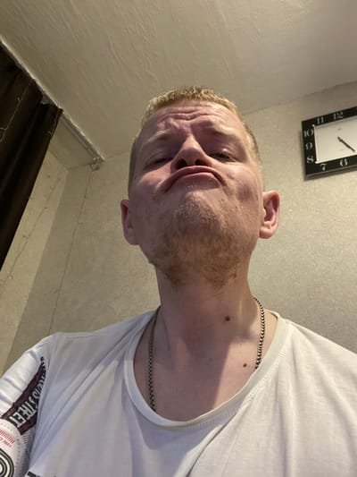 A close-up, low-angle, selfie-style photograph shows a person with blond hair and a light beard making a puckered face. The person is wearing a white t-shirt and a silver chain. In the background, there is a wall with a clock and a dark curtain. The lighting is dim, creating shadows on the person's face and the wall. The environment appears to be indoors, possibly a bedroom or living area.