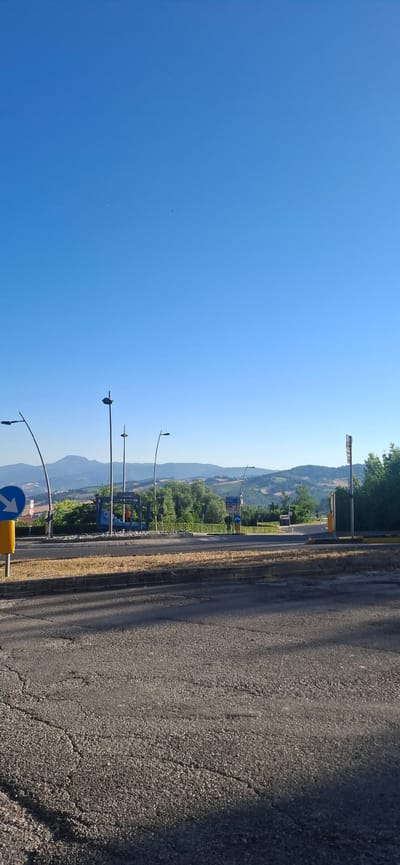 This is a photograph taken on a clear day in Camerino, Italy. The image shows a street scene with a road in the foreground, leading towards a junction with traffic lights and signs. In the background, there are rolling hills covered in trees and vegetation, with mountains in the distance. The sky is a bright, cloudless blue. There are several streetlights visible, as well as a blue bus stop shelter. A yellow pole with a blue directional sign indicating a left turn is prominent on the left side of the image. There are no people visible in the scene. The road surface in the foreground is asphalt and shows signs of wear and cracking. The overall impression is a quiet, rural road on a sunny day.