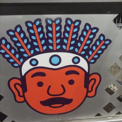 A close-up, front-facing image displays a stylized, flat graphic of a human head, appearing as a sticker or decal, affixed to a grey metallic surface. The head features an orange face with dark purple outlines for the eyes, nose, and a handlebar mustache; the eyes are depicted as small black circles. A white headband adorns the forehead, marked with a central light blue circular accent flanked by two smaller light blue circles. Extending upwards from the head is a stylized headdress composed of eight dark purple, feather-like elements, each outlined in orange and containing multiple small light blue, oval-shaped details. The grey metallic background surface on the right exhibits a pattern of diamond-shaped perforations, through which obscured, blurred background elements are faintly visible. A darker grey, horizontal bar crosses the top edge of the frame, consistent with a structural component or railing. This graphic design, featuring a Betawi-like figure and specific color scheme (orange, purple, blue, white), is characteristic of the branding for the TransJakarta public transportation system, operating within the Special Capital Region of Jakarta, Indonesia.
