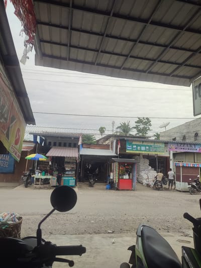 This street-level photograph captures a local commercial area in Kruengmanyang, Indonesia, on an overcast day. The perspective is from under a corrugated metal overhang, with parts of two motorcycles, including a prominent rearview mirror, visible in the foreground.

The scene is dominated by several small, single-story shops or stalls lining a dirt road. In the mid-ground, a shop with a large green banner reads "UD RISKA AHYAR TANI MENYEDIAKAN," indicating it sells agricultural supplies like fertilizer, seeds, and animal feed. The banner also lists an address: "JL. BULUH BLANG ARA - GE KERAMAT - GP KRUENG BANYANG" and a phone number: "0853-1433-8010." Outside this shop, a large pile of white sacks is stacked, and two men are present; one is seated on a motorcycle while the other stands nearby.

To the right of the "UD RISKA AHYAR TANI" shop is another business with a blue sign advertising "AZZAKY COSMETIC PARFUME ACCESORIES" and displaying the numbers "00 20000." To the left of the agricultural shop, a stall operates under a yellow and blue umbrella, with a person visible inside. A red fruit stand is also positioned nearby. Another shop sign, partially obscured, reads "PERDANA CENTER" and shows the "BSI" logo. Overhead, numerous power lines crisscross the sky, and a satellite dish is visible on a distant rooftop. On the far left, a torn banner with the words "PETH MELON" and "ENJOYMENT" is partially visible. The overall impression is that of a bustling, everyday local marketplace or main street.
