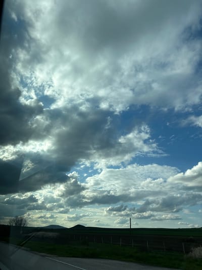 The image presents a view of a sky filled with varying cloud formations, ranging from dense, grey clouds to thinner, lighter ones, against a backdrop of blue sky. The clouds appear to be illuminated by sunlight. Along the horizon, there is a strip of green vegetation and a fence, suggesting a rural or suburban setting. A utility pole is visible against the vegetation. The image is taken from what seems to be an interior perspective, as indicated by a portion of a car or vehicle interior visible at the bottom. While the image captures the sky and landscape, specific information that ties it to the city of Zlatina, Bulgaria, is missing, and the image does not include any people, actions, or interactions.