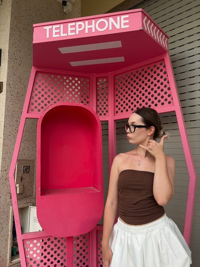 A young woman with dark hair pulled into a bun and wearing glasses stands in front of a large, bright pink, novelty telephone booth. She is wearing a strapless brown top and a white voluminous skirt. Her right hand is raised to her ear in a gesture commonly associated with making a phone call. The telephone booth is decorative and appears to be an art installation or a themed attraction. It has the word "TELEPHONE" in white lettering across the top. The booth is open on one side, revealing a hollow interior. The background consists of a beige, speckled wall on the left and a corrugated metal shutter on the right. The scene is outdoors, possibly on a street or plaza in Nghĩa Trụ Commune, Vietnam.