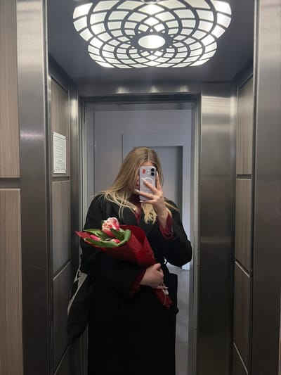 A blonde-haired individual is depicted taking a mirror selfie inside an elevator in Новогорелово, Russia. The person wears a dark, long coat over a red ribbed top and holds a bouquet of pink and white tulips wrapped in red paper in their left arm. A light-colored smartphone, likely an iPhone with a visible MagSafe ring and a small red emblem on its case, is held in the individual's right hand, positioned to capture the reflection. The elevator interior features vertically paneled walls; the left wall has dark wood-grain panels with a vertical strip, and the right wall consists of brushed metal. A decorative, circular light fixture with an illuminated geometric pattern is recessed into the ceiling. A white rectangular sign with text is visible on the left wall panel. The elevator doors behind the individual are open, revealing a light-colored hallway beyond.