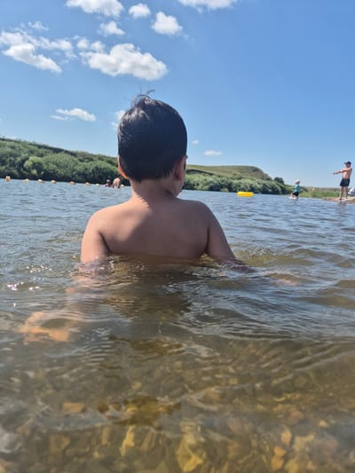 The image shows a child standing in shallow water, facing away from the camera. The child has dark hair and is partially submerged in the water, with their shoulders and back visible. The water is clear with sunlight reflecting off the surface, and the bottom is visible, showing rocks and pebbles.
In the background, there are other people in the water and on the shore. One person is standing on a sandy bank, and another person is further in the water. There is a line of yellow buoys marking an area in the water. Beyond the water, there is a green, grassy hillside under a bright blue sky with scattered white clouds. The overall scene suggests a warm, sunny day at a lake or river. The time of day appears to be midday or afternoon, given the brightness of the sunlight. The location is identified as Новоишим ауылдық округі, Kazakhstan. No text is visible in the image. The atmosphere seems relaxed and recreational, as people are enjoying the water.