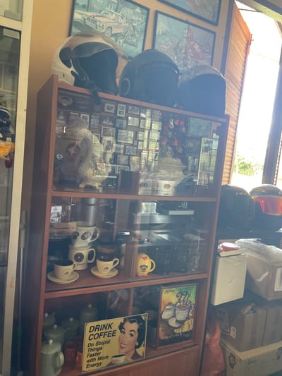 An indoor scene in Ban Yid, Thailand, focuses on a brown wooden display cabinet filled with various items, flanked by a brightly lit window on the right.

On top of the cabinet, three motorcycle helmets are displayed: a white open-face helmet with a clear visor on the left, and two dark-colored full-face helmets in the center and right. Above the cabinet, two framed posters are mounted on a light orange wall; the left poster depicts a vintage car and a motorcycle, while the right poster features a red-clad figure on a motorcycle.

The glass-fronted display cabinet contains multiple shelves:
- The top shelf holds numerous small framed photographs or memorabilia, with a white, ornate decorative item centrally placed. Reflections are visible on the glass.
- The second shelf displays a silver-toned electronic device, possibly an audio receiver.
- The third shelf features multiple cream-colored coffee mugs and saucers, each with a brown graphic, and a yellow ceramic mug on the right.
- The bottom shelf contains various green ceramic containers, including a teapot on the far left. A prominent yellow retro-style sign on the right reads "DRINK COFFEE Do Stupid Things Faster with More Energy" and depicts a smiling woman with a coffee cup. To its left, a framed picture titled "Americano" illustrates a cup of coffee and a croissant.

To the right of the cabinet, a window with wooden blinds allows bright natural light into the room, revealing blurred green foliage outside. Below the window, two additional motorcycle helmets are visible: a black HJC helmet and a black helmet with a reflective orange/red visor. Stacked cardboard boxes are on the floor beneath them, topped by a white rectangular object with a red lid.

To the left of the cabinet, a white vertical partition or door frame is present, with a partial view of a vertical cleaning tool.