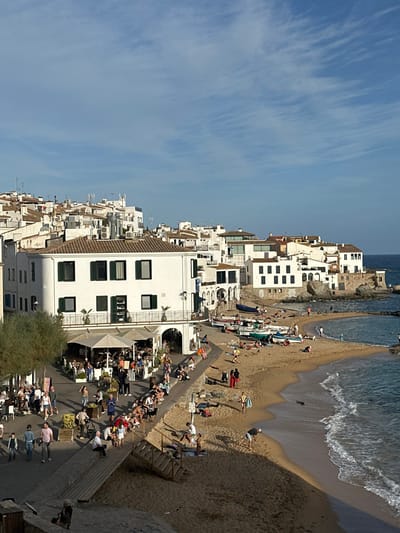 This image captures a panoramic view of a coastal village in Calella De Palafrugell, Spain, under clear, sunlit conditions. The scene is dominated by a cluster of white-washed, multi-story buildings with dark tiled roofs, ascending a gentle hillside from the shoreline.
In the foreground and midground, a paved promenade transitions into a wooden boardwalk, separating the buildings from a crescent-shaped sandy beach. A prominent building with a curved corner, dark green shutters, and a lower-level veranda supporting an outdoor restaurant occupies the left-center. This restaurant features numerous tables, chairs, and large beige or white parasols, under which approximately two dozen patrons are seated and interacting.
The promenade and boardwalk are populated by numerous individuals, estimated at 30-40, casually dressed, walking, standing, and observing the scene. A wooden staircase descends from the boardwalk directly onto the beach.
The sandy beach, extending along the right side of the image, is occupied by approximately 20-30 individuals engaged in leisure activities. Several small boats, predominantly white, blue, and green, are pulled onto the sand. People are seen sunbathing on towels, walking along the water's edge, and one individual appears to be bending over in the sand. The water is a clear blue-green, with small waves breaking gently on the shore. Rock formations are visible at the far right edge, marking the end of the beach.
The sky is a pale blue with scattered, wispy cirrus clouds. The bright sun, likely late afternoon or early morning, casts distinct shadows across the promenade, beach, and parts of the buildings. The overall impression is one of a vibrant, active Mediterranean coastal town.