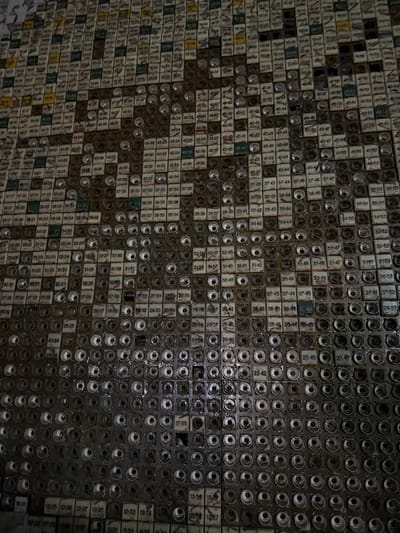 The image depicts a large-scale wall installation, known as the "Wall of Remembrance," located in the National Chornobyl Museum in Kyiv, Ukraine. The installation is composed of thousands of aged, square control panel modules, likely from industrial equipment, arranged in a dense grid pattern. Many modules feature two-digit numerical labels (e.g., "40-40", "35-36", "17-35") printed in various colors. Other modules are characterized by a central circular recess, revealing internal metallic components or a vacant socket, which appear as darker elements. These darker, recessed modules are precisely positioned to form a large, pixelated silhouette of a human head and shoulders in profile, facing left. The modules exhibit visible wear, discoloration, and accumulated grime, consistent with repurposed industrial components. No individuals are present in the frame. The scene is an interior, static display under diffused lighting conditions.