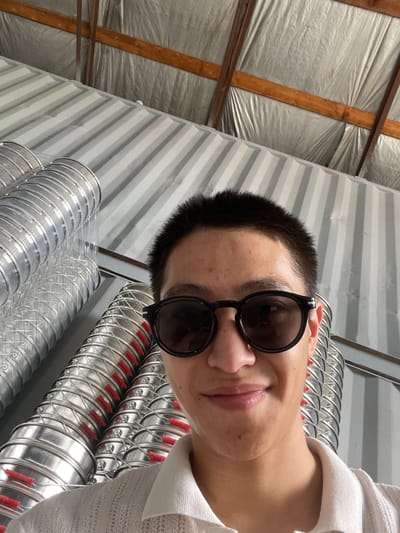 A young East Asian male is taking a selfie in what appears to be a warehouse or industrial setting. He is wearing dark-rimmed sunglasses and a light-colored polo shirt. Behind him, to the left, is a stack of large, corrugated metal pipes, likely for ventilation or ductwork, with red elements visible within their structure. The background consists of a ribbed metal wall and a ceiling with wooden beams and reflective insulation. The overall impression is one of an indoor, functional space, potentially a storage or manufacturing area.