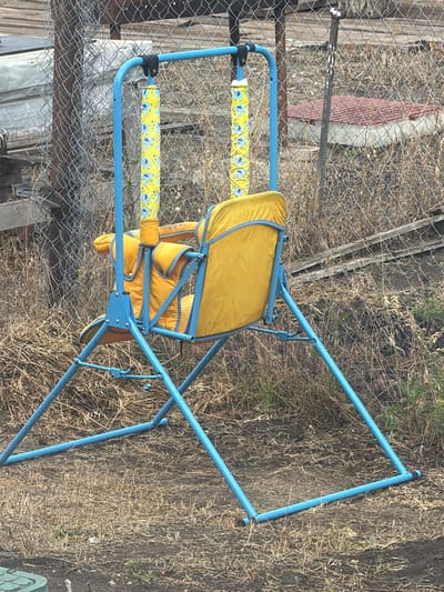 In the city of Krasnyi Krym, Russia, an empty blue metal swing set with a yellow fabric seat and yellow fabric sleeves decorated with blue elephants stands on dry, brown grass and dirt. The swing set is partially obscured by a chain-link fence. In the background, stacked lumber and a red-tiled structure are visible behind the fence. The scene appears to be outdoors in a dry, unkempt area.