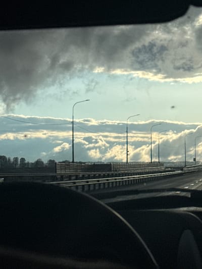 The image captures a view from inside a vehicle, looking out onto a long bridge or elevated road structure under a dramatic sky. The scene is framed by dark, out-of-focus elements of the vehicle's interior on the left and right, suggesting a window or dashboard.
The main subject is a multi-lane roadway or bridge that extends into the distance, appearing on the right side of the frame. Parallel to this road, on the left, are what appear to be railway tracks with ties and rails. A row of streetlights with distinctive curved arms lines the left side of the road/railway structure, receding into the background. Below the tracks, a distant line of trees or land is visible.
The sky occupies a significant portion of the image, dominated by an impressive formation of clouds. A prominent horizontal band of bright, sunlit white clouds stretches across the mid-section, contrasting with darker, grayer clouds above and below it. The light suggests either early morning or late afternoon, with the sun low on the horizon, illuminating the edges of the clouds. A small, blurry reddish dot is faintly visible in the sky in some areas, possibly a distant object or a reflection.
No people are visible. The activity depicted is traveling, likely across a bridge or elevated highway, in a moving vehicle. The weather appears partly cloudy with clear visibility. There is no visible text in the image. The location is contextualized as Aleksandrovskoe, Russia.
