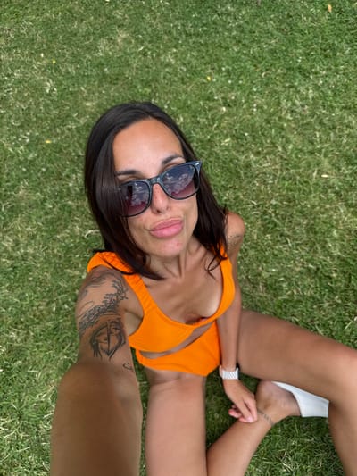 A young woman with dark hair, wearing sunglasses and an orange bikini, sits on a grassy lawn. She is holding a phone in her right hand, seemingly taking a selfie. Her left arm is visible, adorned with tattoos. Her legs are bent, and a white object, possibly a sandal, rests near her right foot. The background is entirely composed of green grass. The image appears to have been taken outdoors, possibly in a park or garden. There are no visible landmarks or structures to suggest a specific location within the city of Grivitsa, Bulgaria.