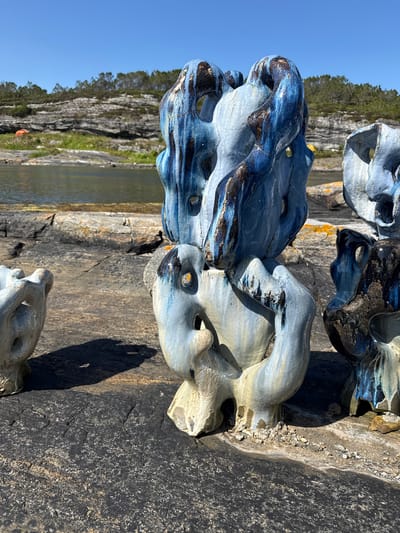 The image depicts an outdoor scene in the city of Rong, Norway, featuring several abstract ceramic sculptures. The sculptures are predominantly blue and light gray, with accents of dark brown and black glaze that appears to be dripping. They have a rough, porous texture and various openings, giving them an organic, somewhat weathered appearance. The sculptures are positioned on a dark, rocky surface, which appears to be part of the natural shoreline. In the background, there is calm water, a rocky incline with sparse vegetation, and a clear blue sky. To the left of the main sculptures, a smaller, lighter gray sculpture is partially visible. The lighting suggests a sunny day, casting shadows from the sculptures onto the rocky ground. There are no people or animals visible in the scene.