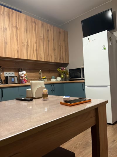 The image captures a portion of a kitchen with a wooden floor and cabinetry. In the foreground, a wooden table with a light-colored countertop is visible. On the countertop, an orange-cased smartphone and a pair of white wireless earbuds are placed near a dispenser of toothpicks. In the background, a large white refrigerator is positioned next to a dark-colored television mounted on the wall. Below the refrigerator, kitchen cabinets with a blue finish and wooden elements are present, along with a microwave and a sink area. A bouquet of flowers sits on a shelf near the sink. The overall scene depicts an indoor domestic setting, likely a kitchen in an apartment or house. There is no indication of the time of day or weather conditions. No people are visible in the image. No readable text is discernible in the image. The location context provided is Редант, Russia.