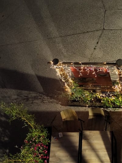 The image shows an outdoor scene at night. A weathered, wooden sign adorned with fairy lights hangs on a post near a stone wall. To the right, there are wooden tables and chairs, with flowers planted nearby. The shadows cast by the tables, sign, and plants stretch across the ground. The lighting is dim, with the fairy lights and a distant light source providing the primary illumination.