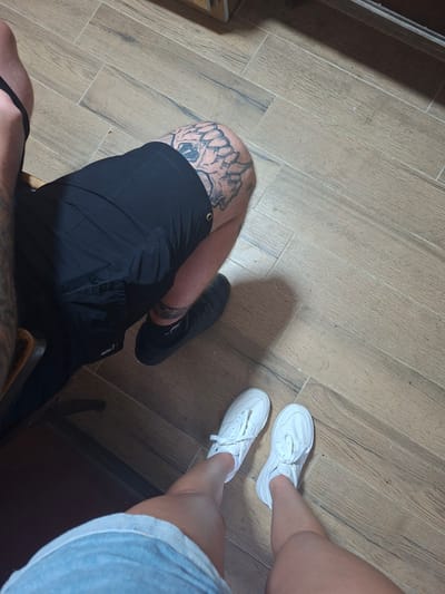 This media file features two individuals from the knees down, standing on a wooden-tiled floor. One person, on the left, is wearing black shorts and a black shoe, with a tattoo visible on their thigh. The other person, on the right, is wearing white sneakers and a light blue denim skirt. The setting appears to be indoors, possibly in a home or public space in Yasen, Bulgaria. The lighting suggests it is daytime, though no specific time can be determined. The image focuses on the lower bodies and footwear of the individuals, with no clear indication of the activity or event taking place beyond them standing.
