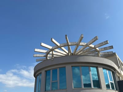 The image displays the upper portion of a building in Razhdak, Bulgaria, set against a clear blue sky with sparse white cumulus clouds. The building exhibits a light gray, textured, curved facade on its upper level, featuring multiple large, rectangular windows with a reflective blue tint that mirror the sky and distant topographical features. Crowning this curved section is a distinctive architectural element: a radial arrangement of light gray concrete beams forming a sunburst pattern. A rectilinear extension of the building, also in light gray, is visible on the far right. No individuals, specific actions, or interactions are present within the scene.
