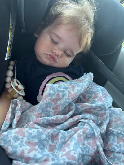 A close-up shot shows a young child asleep in a car seat. The child has fair hair and is wearing a dark blue shirt with a rainbow design on the chest. A patterned blanket, featuring butterflies in pink and blue, is draped over the child. A pacifier with a light-colored ring is visible, and a string of wooden beads is also present. The child's face is peaceful, with eyes closed and lips slightly parted. The setting appears to be inside a car, indicated by the car seat and a seatbelt. The lighting suggests it is daytime. There is a yellow and white sticker with icons on the car seat, but the text is not legible. The location is Lhéry, France.
