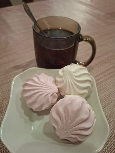 A close-up, eye-level shot shows a white, scalloped-edge plate with three colorful marshmallows placed on it. Two of the marshmallows are light pink, and one is white. A glass mug, filled with a dark liquid, is positioned behind the plate. A spoon rests inside the mug, its handle angled upwards. The mug and plate are set on a textured, pinkish-brown surface, possibly a tablecloth or placemat. The background is softly blurred, indicating a shallow depth of field. The lighting is soft and natural, creating subtle shadows and highlights on the objects. The scene is static and appears to be staged for a photograph. The image was captured in Gorkovskiy, Russia.