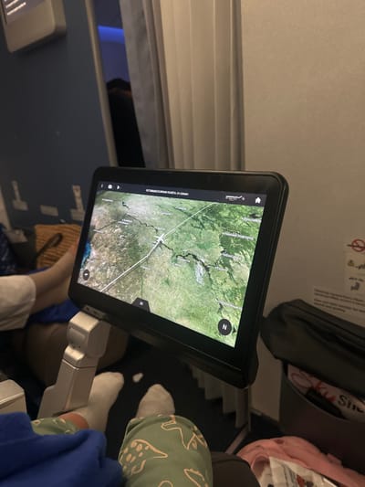 An airplane in-flight entertainment screen displays a satellite image map. The plane's current location is marked. Passengers are visible in the frame, including feet and legs. Overhead compartment, airplane windows, and seat details are also visible. The screen shows a navigation app, potentially tracking the flight path.