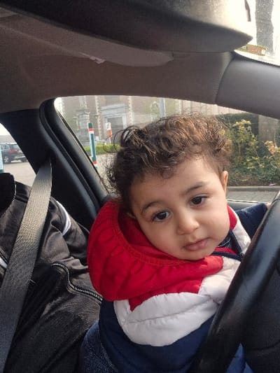 The image depicts the interior of a vehicle in Melle, Belgium, focusing on a young child in the front passenger seat. The child, a toddler with dark, curly hair and dark eyes, wears a red, white, and navy blue hooded puffer jacket and looks directly at the camera with a neutral expression. A black steering wheel is visible on the right, partially obscuring the child. To the left, an adult's torso is visible, wearing a black jacket with white arm stripes and secured by a grey seatbelt. Through the car's window, an overcast or hazy outdoor scene is visible. This includes a multi-story brick building with stone architectural elements in the mid-ground, two striped bollards (one green/white/red, one orange/white), and a dark SUV parked behind them. Green foliage is visible on the right side of the exterior view. The overall environment suggests a daytime setting.