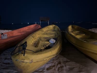 This media file depicts a nighttime scene on a beach, likely in Legonipah, Indonesia. The primary subjects are three kayaks resting on the sandy shore. Two kayaks are prominently in the foreground: one is bright orange and partially visible on the left, while another is yellow and centrally positioned. A third yellow kayak is visible to the right, partially obscured.
The kayaks are the main objects, with one in the center showing a white object inside its cockpit and a dark rope tied to its bow. The scene is set against a dark, clear night sky, with the ocean stretching out to the horizon. Several small lights are visible in the distance over the water, suggesting other boats or structures. In the background, a thatched-roof structure on stilts stands in the water, a common sight in coastal Indonesian areas.
The time of day is clearly night, and the weather appears calm and clear, with no indication of rain or strong winds. The mood is serene and quiet, typical of a beach at night. The sand shows footprints and indentations, indicating recent activity or simply the natural texture of the beach. No people are visible in the frame. There is no visible text in the image.