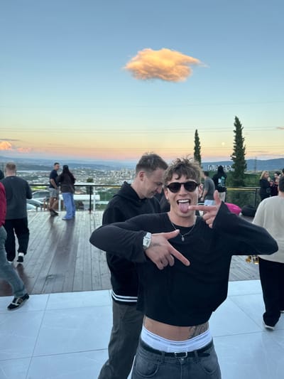 In Mukhatskaro, Georgia, a young man with spiky, light brown hair and sunglasses is the central focus of this outdoor evening scene. He is wearing a black long-sleeved shirt, dark gray jeans, and white boxer briefs. He has a tattoo on his left hip and is pointing his index finger towards his left while sticking his tongue out and making a peace sign with his right hand. He is positioned on a light-colored tiled area, with a wooden deck behind him. Several other individuals are present in the background, some interacting with each other and others standing or walking. The backdrop features a city skyline with mountains in the distance, under a sky with a single orange-tinted cloud and a faint moon. Two tall, dark green trees stand prominently on the right side of the frame.