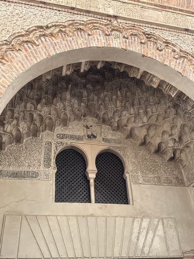 The image shows an ornate archway with intricate Islamic geometric patterns and calligraphy adorning the ceiling. Below the arch, there are two arched windows with decorative latticework. The architecture suggests a historical building with Moorish influences, commonly found in Granada, Spain. The materials appear to be stone and plaster with painted embellishments. The lighting is natural, casting subtle shadows that highlight the detailed carvings. There are no people visible in the image.