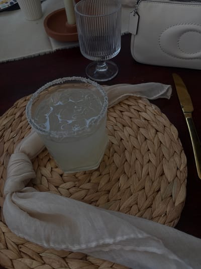 The image shows a table setting, likely indoors, with several objects arranged on a dark wooden surface. A handbag, light-colored and potentially made of leather, is positioned on the upper left side. Adjacent to it, a golden knife is placed horizontally. A glass with a salted rim containing a beverage sits on a woven placemat. A loosely knotted light-colored cloth napkin partially covers the placemat and extends towards the glass. Another textured glass is lying down next to a small clay pot. The image doesn't contain sufficient information to confirm the specific location in Santa Cruz Tehuixpango, Mexico.