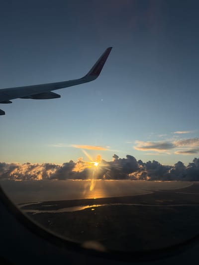 An aerial view from an airplane window captures a sunset over a body of water and land. The sun is partially obscured by clouds, casting golden rays across the sky and reflecting on the water's surface. The airplane's wing, with the Wizz Air logo visible, extends from the upper left corner of the frame. The scene is set within the geographical coordinates of Czerwony Kościół, Poland.