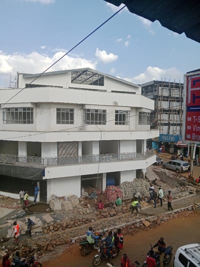 The scene is a partially constructed commercial building in Kapsabet, Kenya, located at latitude 0.202 and longitude 35.098. A large, white, three-story building with a partially constructed roof and a balcony on the second floor sits on a dirt lot. Several construction workers are working on the lot in front of the building, laying bricks and moving rocks. There are several other buildings in the background, one of which is also under construction and has scaffolding on it. There are a few vehicles parked on the street in front of the building, including a white pickup truck and a silver car. There is a sign advertising TECNO mobile phones. The sky is partly cloudy.