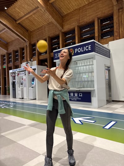 A lone female is depicted mid-action, appearing to juggle or throw a bright yellow ball inside a public service building located in Tianya District, Sanya City, China. The woman, with fair skin and dark hair braided over her left shoulder, wears a light beige short-sleeved t-shirt, dark gray athletic leggings, a light teal long-sleeved top tied around her waist, and gray and black hiking boots. She stands facing slightly right, smiling broadly with her head tilted back, and both hands are open and raised towards the bright yellow ball suspended in the air above her.

In the background, to the right, is a white police service booth with a blue top section displaying "POLICE" in English and Chinese characters. Further signage on the booth identifies it as a "Temporary Driving Permit Foreigner Service Window" in Chinese, English, and Russian. To the left, an "INTELLIGENT SERVICE AREA" sign in English and Chinese is visible in front of several white self-service kiosks. The interior features a high, vaulted ceiling constructed from warm-toned wooden beams and slats. The upper walls are paneled with dark wood and incorporate rectangular window-like structures with frosted or illuminated panels. The floor is tiled in a pattern of light grey and white squares, overlaid with a prominent blue and green striped pathway featuring large white directional arrows pointing to the right.