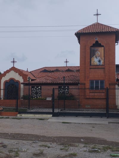 The image shows a church-like structure in Barquisimeto, Venezuela. The building is constructed primarily of red brick with a red tile roof. The front of the property is enclosed by a black metal fence. On the right side of the structure is a tower-like extension adorned with a portrait of a religious figure. The building has crosses on the roof and a distinctive arched doorway. The ground in front of the fence appears to be a dirt road or unpaved area. The sky is overcast.