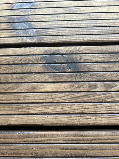 This is a close-up, high-angle shot of a weathered wooden surface, likely a deck or fence, composed of vertical wooden planks. The wood grain is prominent, showing a natural, textured pattern with variations in color ranging from light brown to dark, almost black hues. Several knot formations are visible within the planks, some appearing as dark, circular patterns, while others are more subtle. There are also signs of age and wear, such as faint cracks and a rougher texture in some areas, particularly around the knots. The lighting suggests it is daytime, casting subtle shadows that emphasize the grooves between the planks and the dimensionality of the wood grain. The overall impression is one of natural, rustic material, likely exposed to the elements over time. The scene is devoid of people or any discernible objects beyond the wooden structure itself, focusing entirely on the texture and pattern of the wood. The location context of Haaslava, Estonia, suggests this could be an outdoor setting in a rural or suburban environment. There is no visible text in the image.