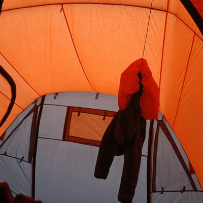 Inside a tent at Gilman's Point, Tanzania, an orange outer tent fabric ceiling is visible, speckled with numerous water droplets, indicating condensation or external moisture. Red structural seams are present on this orange fabric. Below, a grey inner tent wall features a rectangular window or vent with an orange-brown transparent or mesh panel. Suspended from a thin black cord hanging from the tent ceiling are two garments: an upper, bright orange, puffy hooded jacket, and a lower, darker brown or black garment, possibly a pair of trousers or a second jacket, both hanging vertically. A dark-clad hand or arm is partially visible in the bottom left corner of the frame. The scene is static, devoid of active human interaction, focusing on the interior conditions of the tent.