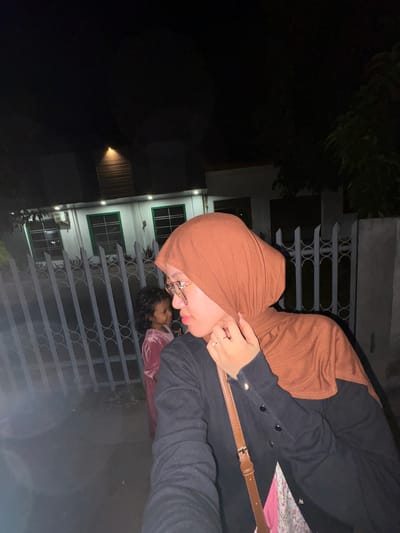 A woman wearing a brown hijab and glasses is in the foreground of this nighttime outdoor image, her head turned to the left and her right hand touching her hijab. She is wearing a black cardigan over a floral-patterned garment. A brown crossbody bag is slung over her shoulder. In the middle ground, a young girl in a pink dress stands behind a decorative metal fence. Behind the fence is a building with green-trimmed windows and an air conditioning unit. The scene appears to be in the city of Tanggul, Indonesia, as indicated by the metadata.