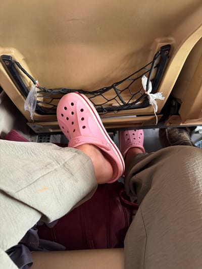 The image shows a close-up, first-person perspective of a person's legs and feet, wearing pink Crocs and light-colored pants, resting in a vehicle's passenger seat. The backdrop reveals the interior of a car, specifically the back of a front seat with a mesh pocket. A maroon bag is visible beneath the person's legs. The environment suggests a travel or commute context, likely within the city of Phakhao, Laos, given the inferred location.