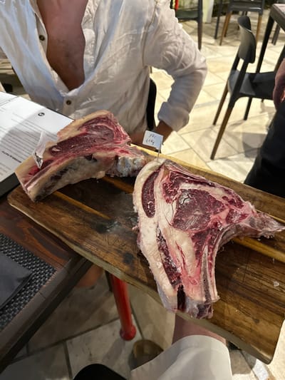 An indoor scene in a restaurant in Nardò, Italy, displays two large cuts of raw, bone-in meat presented on a rustic wooden cutting board. The meat on the right features a white tag pierced by a metal skewer, bearing the handwritten inscription "X01 4.4 Kg," indicating its weight. The cutting board rests on a dark wooden table with a dark, textured placemat visible on the left.

In the upper left quadrant, a person is seated at the table, wearing a light-colored linen shirt with an open collar, their left arm visible beside an open menu or document. The lower right quadrant shows a leg and foot, clad in a light-colored shoe, beneath the table. The background reveals light-colored, possibly tiled, flooring and dark, modern-style chairs, consistent with a dining establishment. Partial views of other tables, including one with a red support leg, are also present.