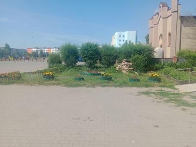 The image depicts an outdoor scene on a sunny day, likely in the city of Atbasar, Kazakhstan, given the context of the provided crops. In the foreground, a paved area of interlocking gray bricks is visible, with patches of sparse grass and weeds growing in between. A low strip of green grass runs along the middle ground, with several rectangular planters containing yellow and red flowers. Behind the planters, a cluster of small, round, leafy green bushes is present. Further back, a stone retaining wall is visible.
To the right, a pale tan building with Gothic architectural elements, including pointed arches and decorative spires, stands prominently. Adjacent to it, a section of wooden fencing is seen. Behind the bushes and stone wall, a light blue and yellow multi-story apartment building with white-framed windows is visible. Further in the distance, other apartment buildings and structures with red and white facades can be seen, along with a tall flagpole flying a flag. A children's playground with colorful structures is also visible on the left side of the image. A few individuals are scattered throughout the background, walking near the playground and along the paved areas. The sky is clear and blue.