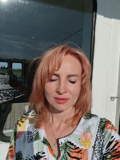 This is a medium close-up, outdoor, portrait-style photograph of a woman with reddish-pink hair. She is positioned in the foreground, with her eyes closed and a serene expression on her face. She is wearing a colorful, patterned shirt with a white lace trim around the collar. A delicate silver necklace with a crown pendant is visible around her neck.
Behind the woman, a large window reflects a scene of a body of water with shimmering light, and what appears to be a treeline in the distance. There are also reflections of windows and the interior of a structure. To the right of the woman, part of a white building with a dark trim is visible, suggesting the photograph was taken on a boat or a structure adjacent to water.
The lighting suggests it is daytime, likely late afternoon given the warm tones of the light on the woman's face. The weather appears to be clear and sunny, with the bright sun casting shadows and highlighting the water's surface. The setting seems to be a calm, possibly rural or natural environment, given the reflection of the treeline. The overall mood is peaceful and contemplative. There is no visible text in the image.