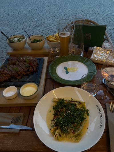 The image depicts a table setting at a restaurant in Sterzing (Vipiteno), Italy. A meal is laid out on a wooden table, with a cobblestone-like patterned surface visible in the background. The scene appears to be outdoors, possibly a patio or terrace.
The main subjects are various dishes and dining implements. On the right, a white plate with a black rim is filled with what appears to be a pasta dish topped with chopped green herbs and resting in a creamy sauce. The plate has the text "SCHWARZER ADLER" written around its rim. To the left of this dish, a dark stone slab holds sliced, cooked meat, possibly steak, garnished with rosemary. Beside the meat are two small white bowls, one containing a creamy white substance (likely a sauce or dip), and the other filled with coarse salt. Further to the left are two more small white bowls, each containing a salad-like mixture, with spoons inside. A white bowl containing French fries is also visible.
Several glasses are present on the table, including two clear stemmed glasses, a glass tumbler with a yellowish liquid, and another small glass. A dark green menu or booklet is propped up against a wicker-like object. A decorative green plate with a hand-painted yellow lemon design is positioned in the center. A fork and knife are neatly placed on a brown napkin on the right side of the table, next to the plate of pasta.
The lighting suggests it could be daytime, with soft lighting that doesn't cast harsh shadows. The overall impression is a well-presented meal, ready to be enjoyed in a pleasant outdoor setting. The text "SCHWARZER ADLER" indicates the name of the restaurant.