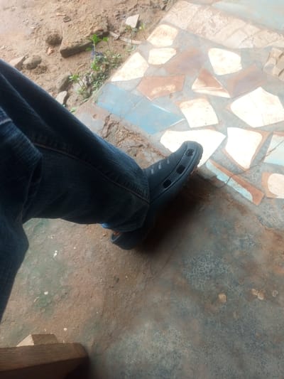 The image shows a person's legs and feet wearing blue jeans and black crocs. The person is sitting on a step, with their left leg crossed over their right. The background is a mosaic tiled floor with brown and blue tiles. There is also some dirt and gravel visible on the ground. The scene appears to be outdoors, possibly in a residential area of Agbara, Nigeria.