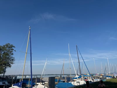 This image depicts a sunny day at a marina in Gstadt am Chiemsee, Germany. The scene is dominated by a clear, bright blue sky with a few wispy clouds. In the foreground, a row of sailboats is moored along a dock. Many of the boats are covered with tarps, suggesting they are either being stored or are awaiting their next use. Some masts are visible, with one prominent mast on a sailboat in the left foreground featuring a yellow spar and a blue sail cover.

In the background, the calm waters of a lake stretch out, with several other boats visible in the distance. The far shore is lined with trees and appears to be a natural landscape. On the right side of the image, two people are seated on a grassy bank overlooking the marina. They appear relaxed, enjoying the pleasant weather. The overall atmosphere is peaceful and recreational. The time of day appears to be late morning or early afternoon, given the bright sunlight and the length of the shadows.