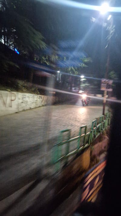 The image depicts a street scene at night in Kullu, India. The road is paved with cobblestones and appears wet, reflecting the light from street lamps and a passing motorcycle. A motorcycle is visible in the middle of the road, its headlight illuminating the wet surface. To the left, a concrete wall has some writing on it, including what appears to be dates and possibly some text in Hindi. There is a green metal railing along the edge of the road on the right. Trees and foliage are visible in the background, suggesting a natural setting within the city. The overall impression is of a dimly lit, possibly foggy or misty, environment.