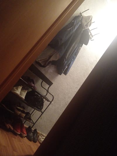The image depicts an interior scene, possibly a hallway or entryway, with a focus on a shoe rack and a coat rack. The lighting is uneven, with a bright light source coming from the upper right, casting long shadows.
On a multi-tiered metal shoe rack, several pairs of shoes are visible. The bottom shelf holds a pair of red and white shoes, followed by a pair of dark boots. Above these, on the next shelf, are two white ankle boots. There is also a dark bag or piece of luggage on this shelf. The topmost shelf is mostly obscured.
Above the shoe rack, a metal coat rack is mounted on the wall. Several coats are hanging on it, including a dark blue jacket with a hood. The wall appears to be textured.
To the left, a wooden doorframe is visible, suggesting the space is likely an entryway. The floor is made of wooden planks. The overall impression is one of a functional storage area.
The location is identified as Shakhtinsk, Kazakhstan. However, there are no specific visual cues in the image that would definitively place it in Shakhtinsk or any particular city. The scene is domestic and does not contain any identifying landmarks or features.