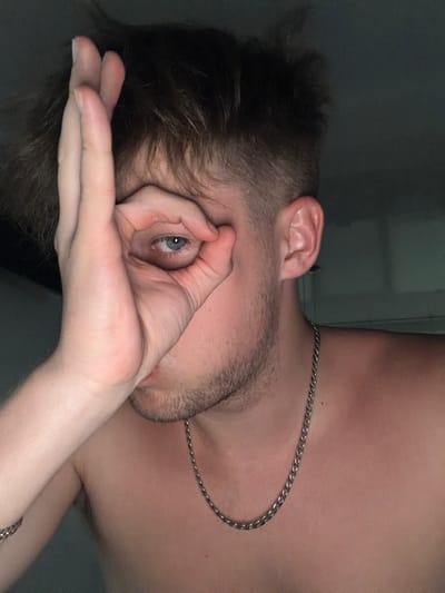 A young man with short, spiky brown hair and blue eyes is shown in a close-up portrait. He is shirtless and wearing a silver chain necklace. His right hand is held up to his face, with his thumb and index finger forming a circle around his eye, as if looking through a lens. The background is dark and nondescript, suggesting an indoor setting. The image does not contain any elements that would indicate its location in Vanino, Russia, or any other specific city.