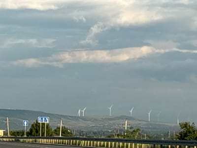 This is a landscape photograph taken from a highway looking towards a rural area with rolling hills under a cloudy sky. In the foreground, a section of the highway barrier is visible, with wooden posts and metal railing. Two blue road signs are mounted on poles to the left of center. The left sign is smaller and shows a single white upward arrow. The sign to its right is larger and displays three white upward arrows. Behind the signs and highway, a dense line of dark green trees separates the immediate foreground from the rolling, grassy hills. Several wind turbines, white with three blades each, are scattered across the tops of these hills, appearing as distant white silhouettes against the overcast sky. The sky itself is a mix of gray and white clouds, with some lighter, brighter patches indicating diffused sunlight. The overall impression is one of a vast, somewhat industrial landscape in the region of Kvemo Akhalsopeli, Georgia, characterized by open terrain and renewable energy infrastructure.